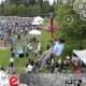 Large corporate fun event image featuring many people on a warm summer's day. A man dangles from a zip line with the crowds below on the grass covered showground