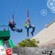 2 men gliding down a zip line, over a green tree while attending a fun corporate event. there's a beautiful clear blue sky behind them.