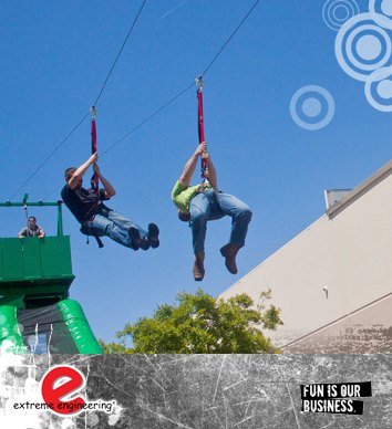 2 men gliding down a zip line, over a green tree while attending a fun corporate event. there's a beautiful clear blue sky behind them.