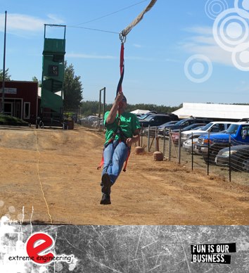 A team member of a mobile zip wire hire company testing the line after the main tower had been erected. The man is gliding over a sandy area with a line of cars to the right of the image.