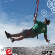 Man in a hat and green t-shirt, gliding down a zip line with a blue sky behind and a few fluffy white clouds